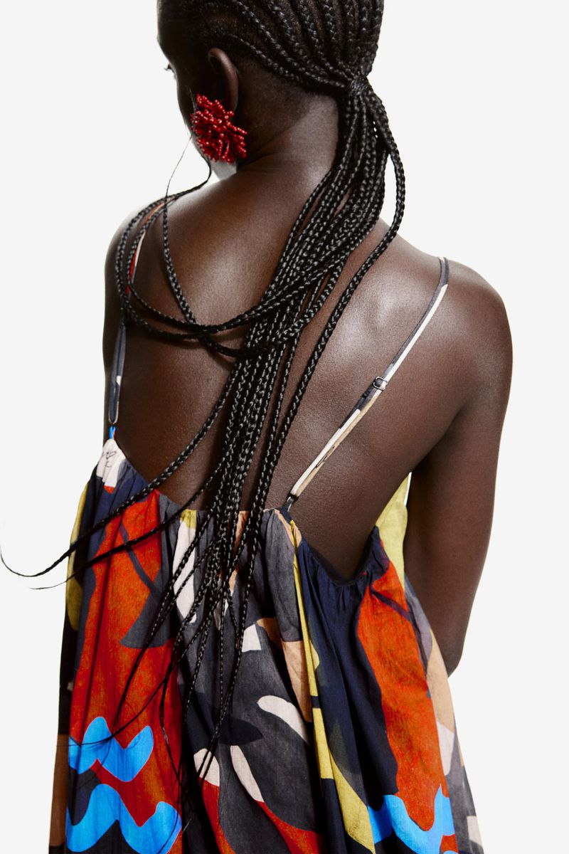 Back view of an African American woman with braided hair, wearing a colorful spaghetti strap dress and red flower earrings on a white background.