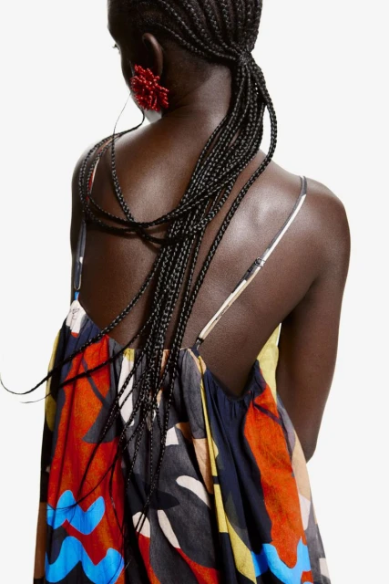 Back view of an African American woman with braided hair, wearing a colorful spaghetti strap dress and red flower earrings on a white background.
