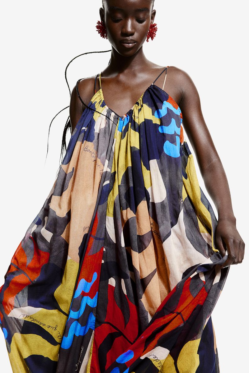 A dark-skinned woman in a vibrant, flowing dress stands against a white background, accessorized with striking red earrings.