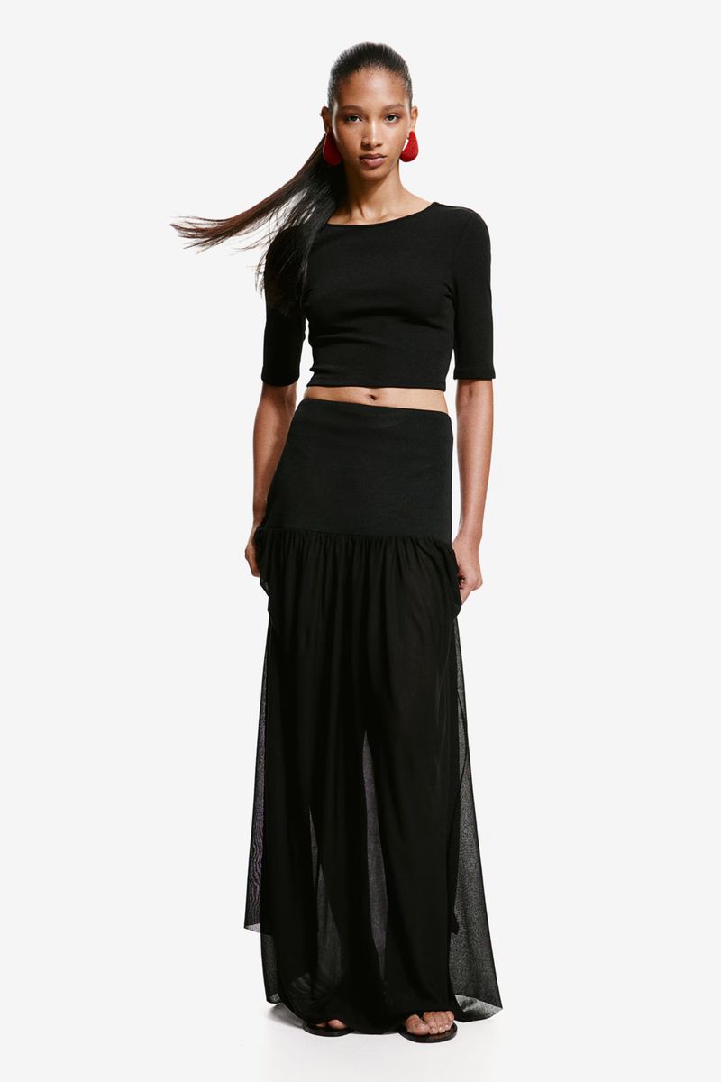 Model wearing a black crop top, dropped-waist sheer maxi skirt, and red statement earrings against a white seamless background.