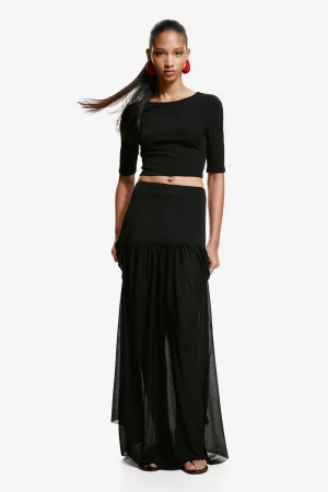 Model wearing a black crop top, dropped-waist sheer maxi skirt, and red statement earrings against a white seamless background.