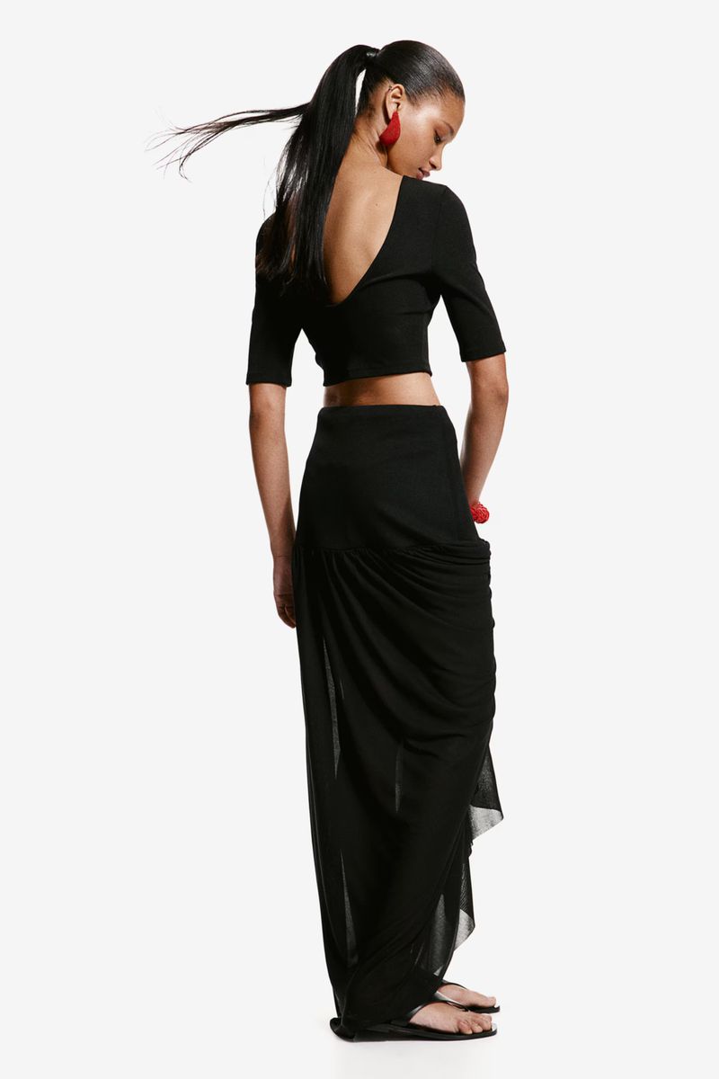 Back view of a young adult female with long black hair in ponytail, wearing a black crop top and skirt, and red earrings, on white.