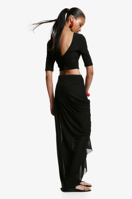 Back view of a young adult female with long black hair in ponytail, wearing a black crop top and skirt, and red earrings, on white.