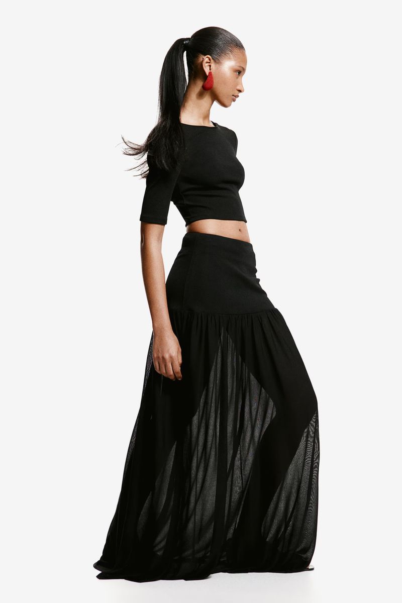 Fashion shot of a slender woman in a black crop top and long black semi-sheer skirt, posing in a studio environment on a white backdrop.