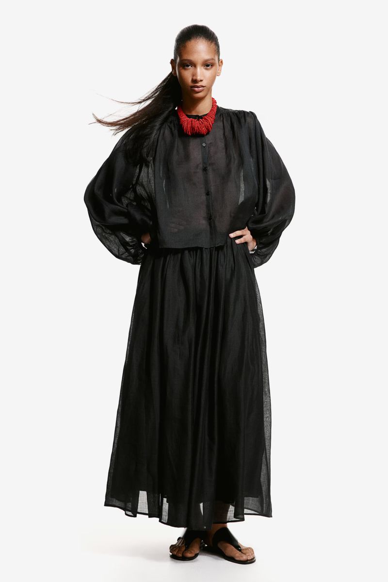 Full-length studio shot of a Black female model in a long black dress and sheer black top with a red necklace, hands on hips.