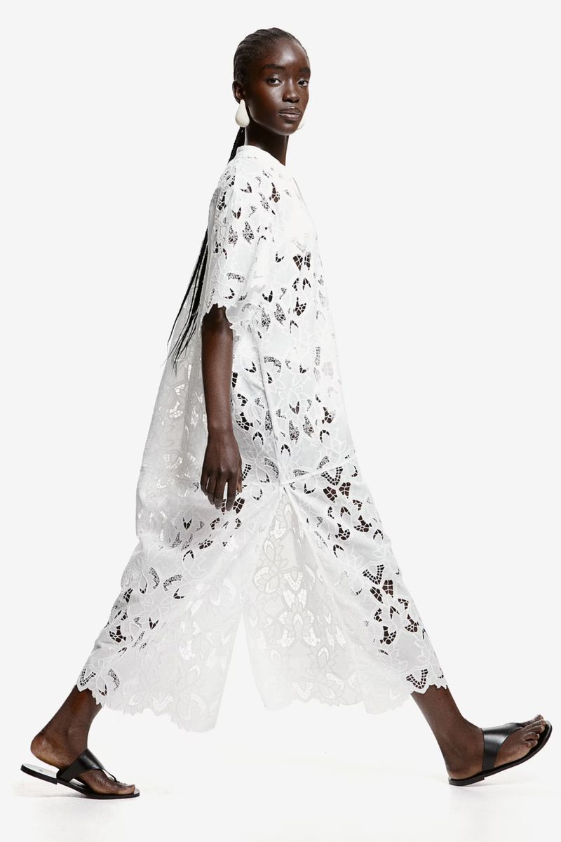 A dark-skinned woman walks in a white patterned dress and black sandals against a white backdrop in a fashion shot with studio lighting.