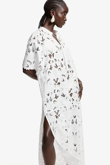Fashionable woman in a white patterned maxi dress with a side slit stands against a bright white background, showcasing modern style.