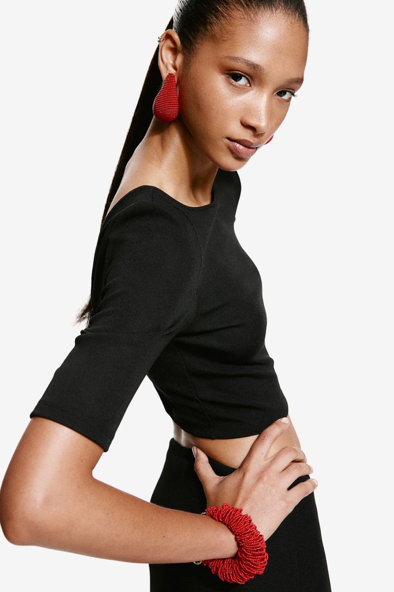 Headshot of woman in black crop top and skirt posing with red bead jewelry against a white backdrop, fashion, studio lighting.