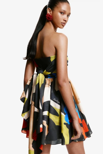 A woman poses in a colorful strapless mini dress with an abstract print, accentuated by large red earrings against a clean white backdrop.