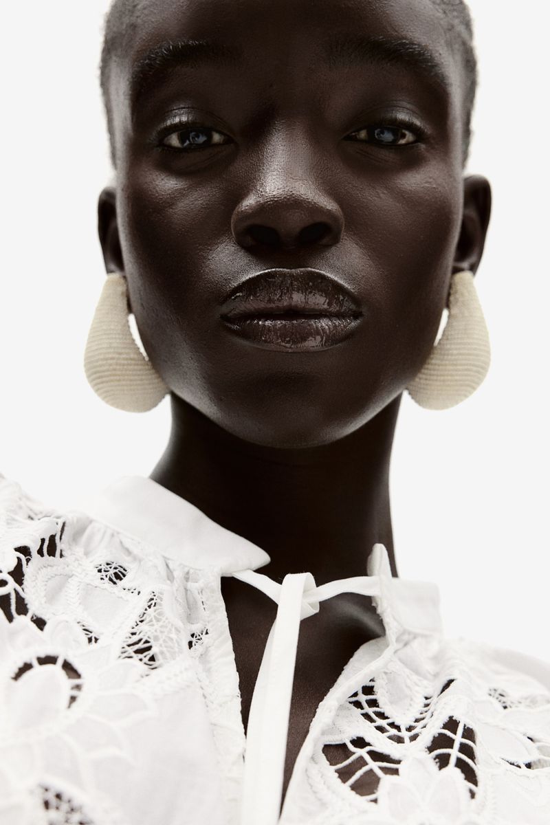Close-up of a woman with dark skin, wearing a lace top and large teardrop earrings, set against a stark white background.