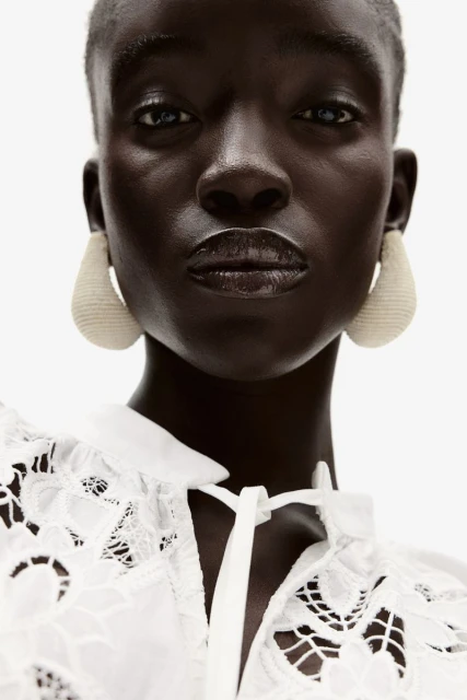 Close-up of a woman with dark skin, wearing a lace top and large teardrop earrings, set against a stark white background.