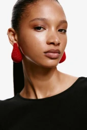 Close-up portrait of model Maty Fall wearing large red beaded statement earrings and a black top against a white studio background.