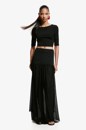 Model wearing a black crop top, dropped-waist sheer maxi skirt, and red statement earrings against a white seamless background.