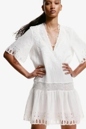 Model posing with hands on hips wearing a white embroidered lace-trimmed mini dress against a seamless white background.