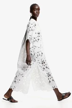 Model Maty Fall in profile wearing a white eyelet lace dress and black flat sandals against a solid white studio background.
