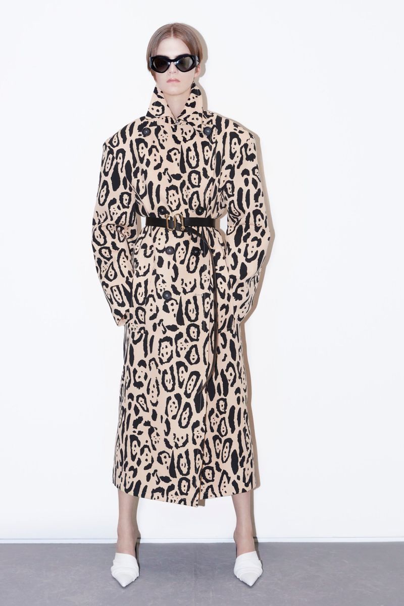 A woman stands in a full shot, wearing a tan animal print coat with a black belt and white heels against a white backdrop.