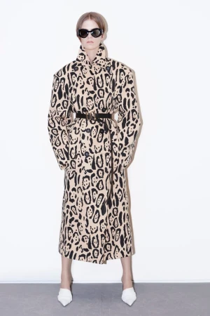 A woman stands in a full shot, wearing a tan animal print coat with a black belt and white heels against a white backdrop.