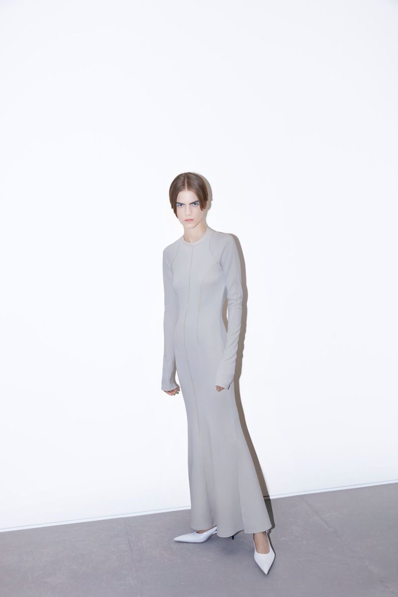 A slender adult female fashion model poses in a long-sleeved fitted light-grey dress and white pointed heels against a bright white backdrop.