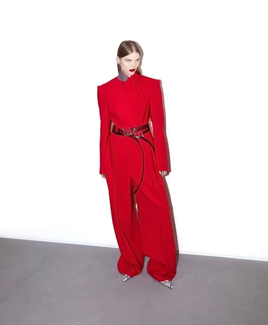 A fair-skinned woman poses in a red pantsuit with a statement belt against a white backdrop in a fashion forward studio shot.