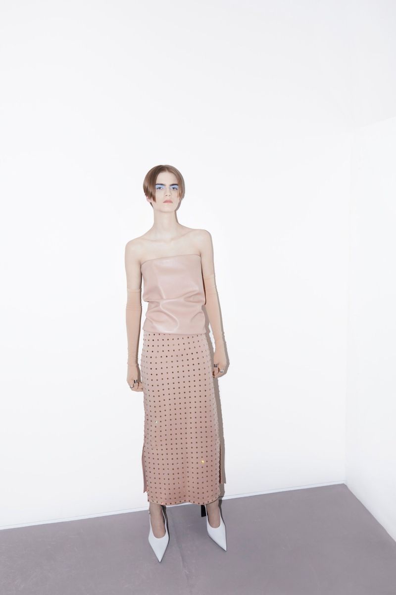 A slender woman with short hair poses in a light pink top and embellished skirt, paired with white heels, in a minimalist studio setting.