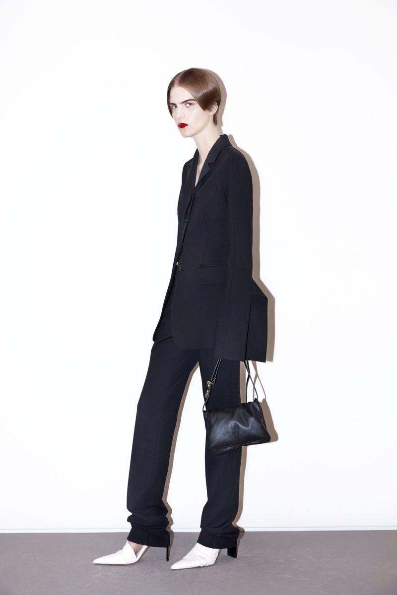 Model wearing a black tailored pantsuit with flared cuffs and white pointed-toe heels against a white studio wall.