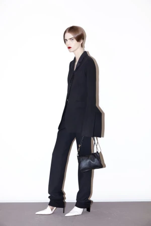 Model wearing a black tailored pantsuit with flared cuffs and white pointed-toe heels against a white studio wall.