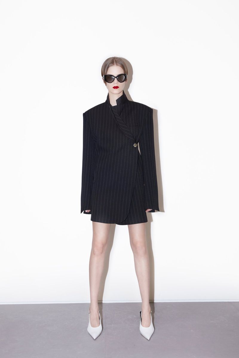 A woman models a pinstripe blazer dress with white heels and sunglasses against a stark white backdrop in a fashion-forward pose.