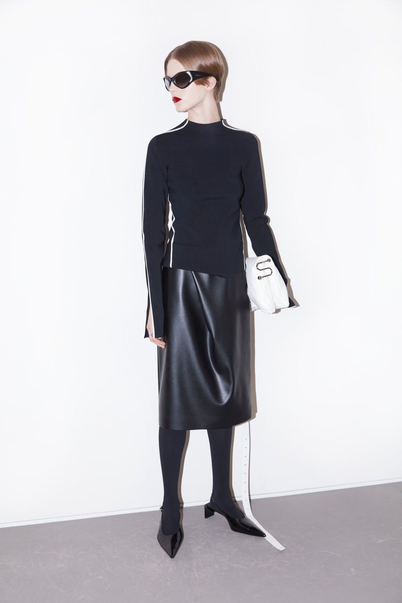 A woman wearing a black top, leather skirt, tights, and shoes is standing in a white photo studio with a purse, looking elegant.