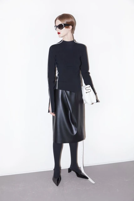 A woman wearing a black top, leather skirt, tights, and shoes is standing in a white photo studio with a purse, looking elegant.