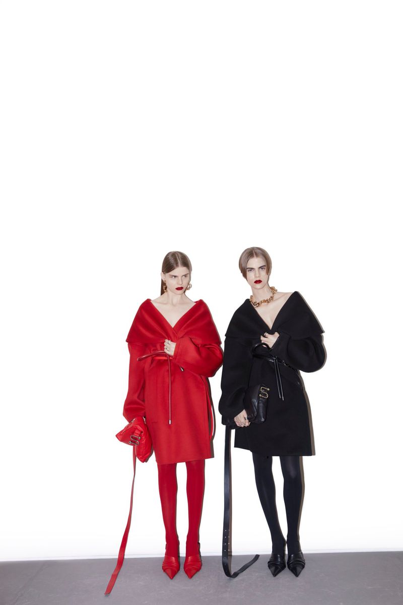 Two women pose in matching fashion coats and leggings, one in red with a matching purse, the other in black with a black purse.