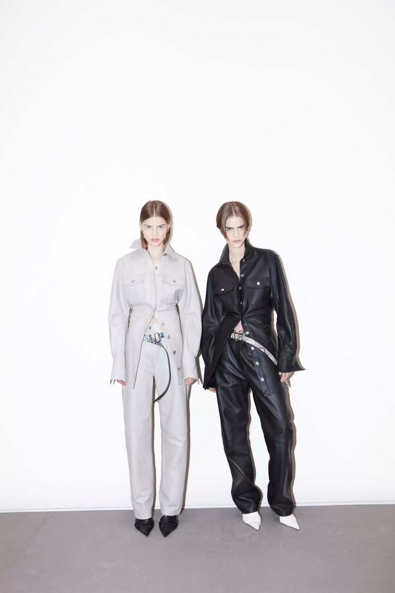 Two fashion models pose in matching leather outfits against a white background, creating a striking studio portrait with a minimalist aesthetic.
