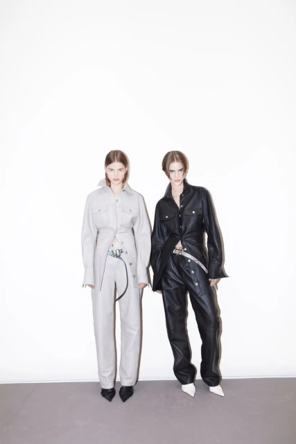 Two fashion models pose in matching leather outfits against a white background, creating a striking studio portrait with a minimalist aesthetic.