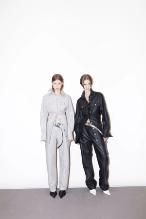 Two fashion models pose in matching leather outfits against a white background, creating a striking studio portrait with a minimalist aesthetic.