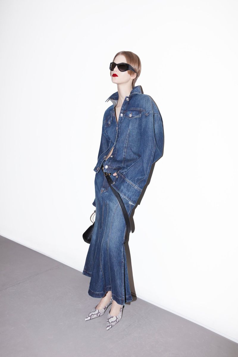 A fair-skinned adult female fashion model in a denim outfit and snakeskin print heels poses in front of a white wall in a photo studio.