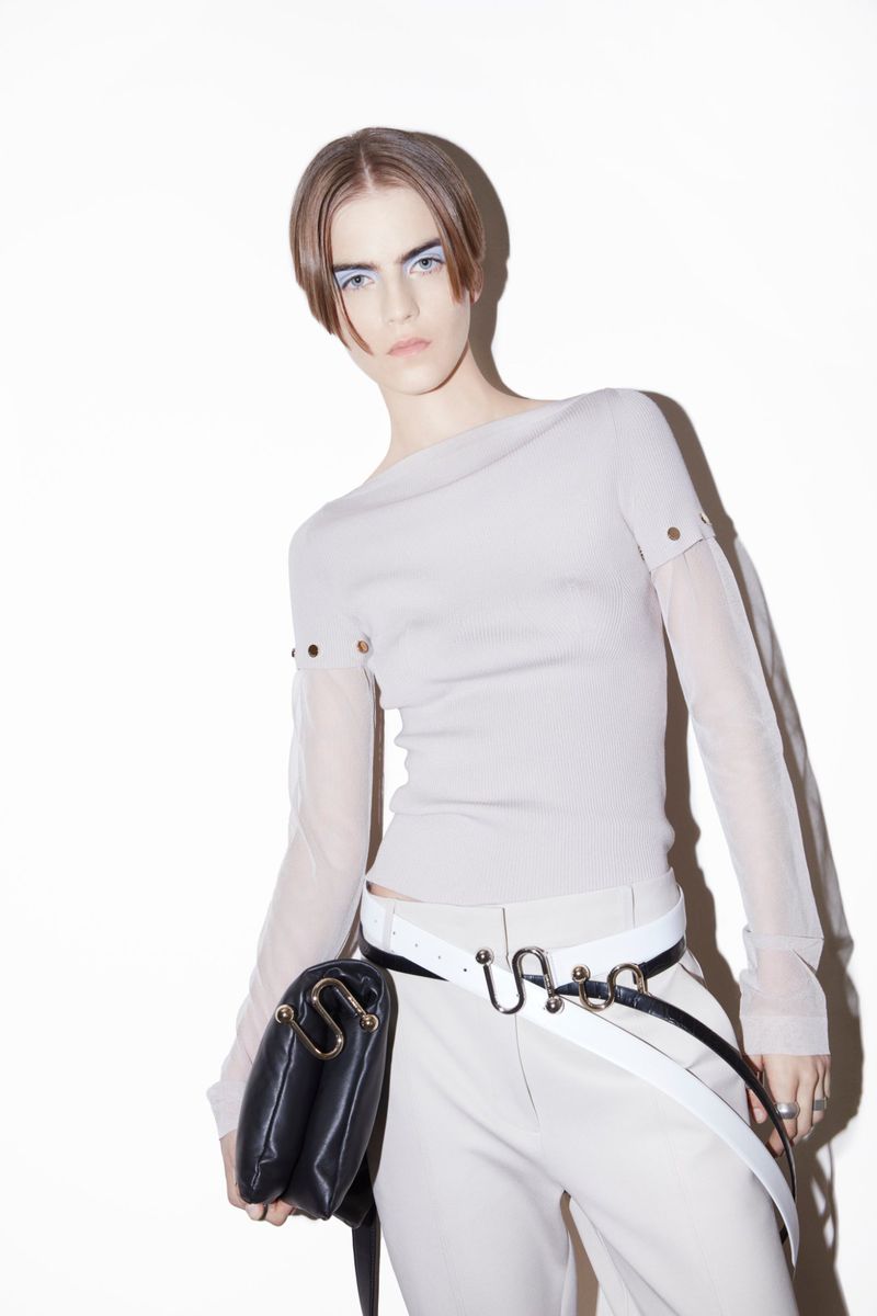 A fashion shot of a model in a neutral top, light trousers, and black purse on a white background, posing in a studio setting.