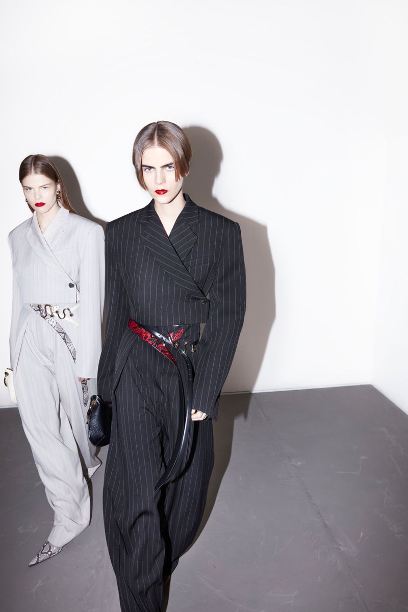 Two female fashion models in pinstripe suits, one black, one grey, pose indoors in a high-fashion editorial style against a stark white wall.