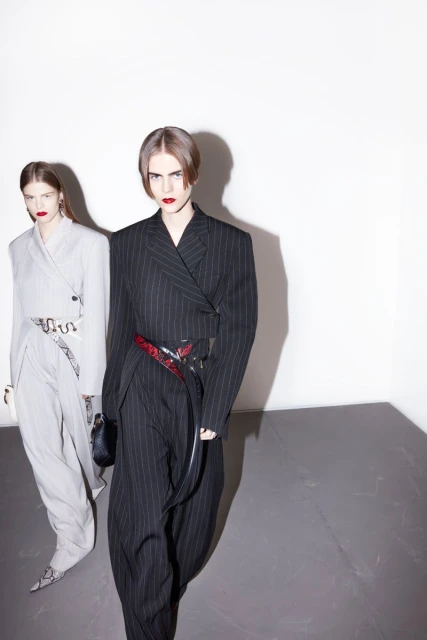 Two female fashion models in pinstripe suits, one black, one grey, pose indoors in a high-fashion editorial style against a stark white wall.
