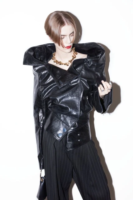 Adult woman in a studio wearing an oversized black leather jacket, pinstripe pants, and a gold necklace, looking down at the camera.