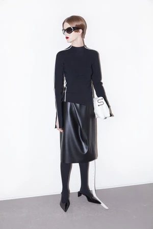 Model posing in a black long-sleeve top, black leather midi skirt, black pointed slingback heels, sunglasses, and holding a white bag.