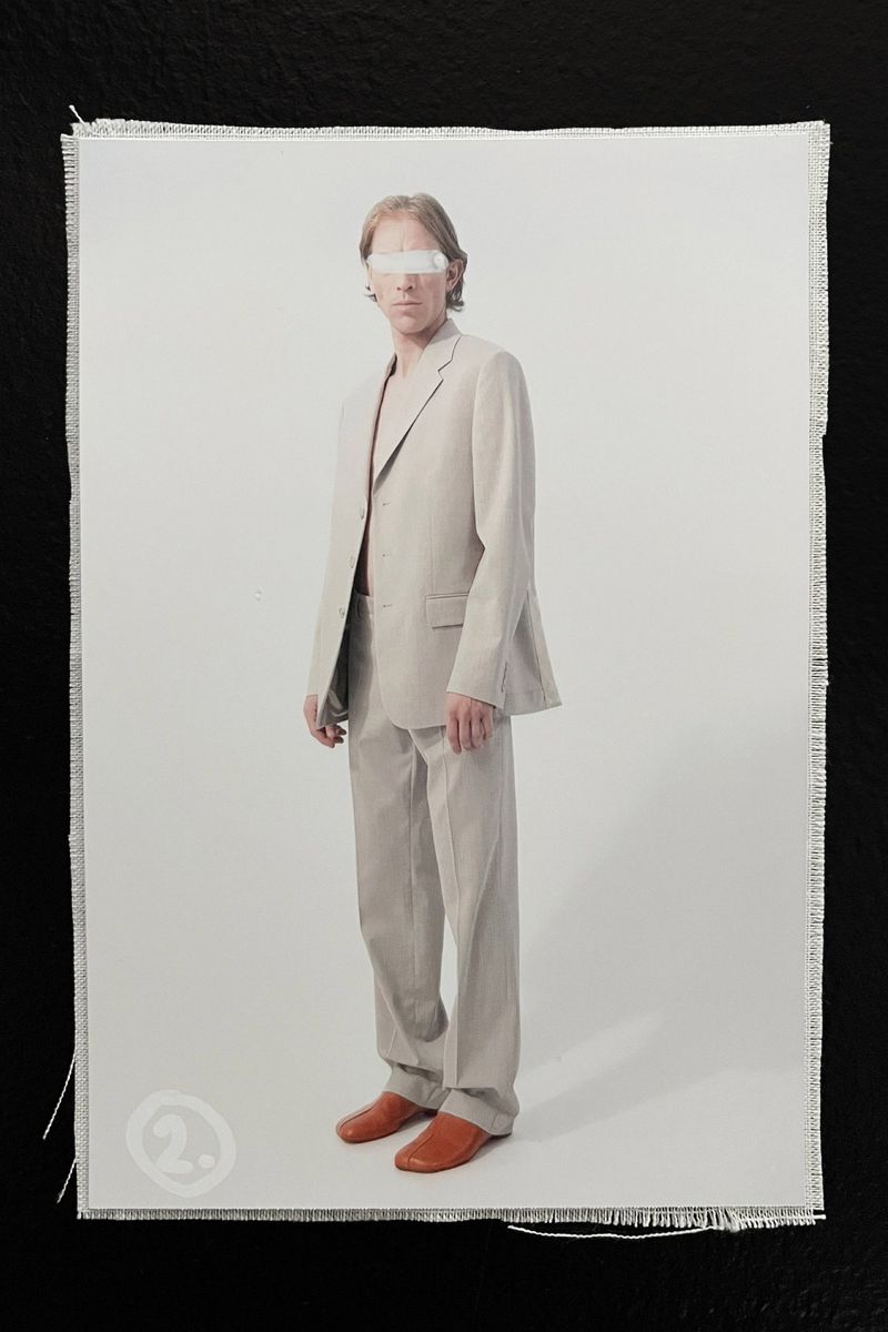 Adult male model in a grey suit and orange loafers standing indoors in front of a white background on a fringed canvas.