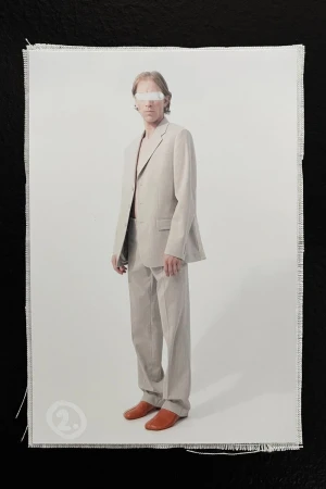 Adult male model in a grey suit and orange loafers standing indoors in front of a white background on a fringed canvas.
