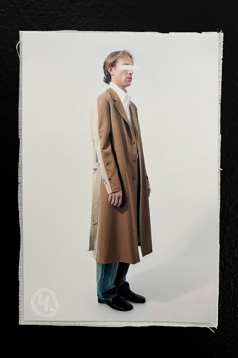 Full-length studio shot: adult male poses in a tan trench coat over a white shirt and blue jeans, on off-white against a black backdrop.