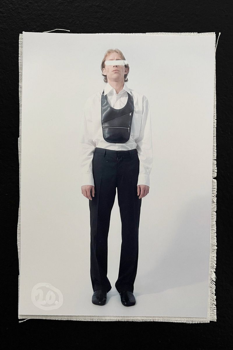 Full-length studio shot of androgynous adult in a white shirt, black trousers, shoes, and a leather crossbody bag on a white background.