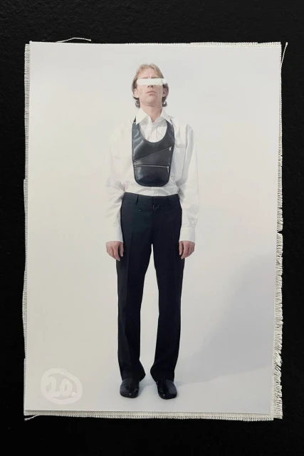 Full-length studio shot of androgynous adult in a white shirt, black trousers, shoes, and a leather crossbody bag on a white background.