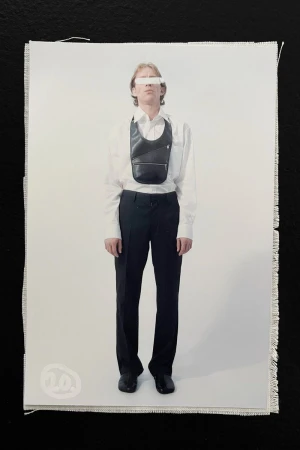 Full-length studio shot of androgynous adult in a white shirt, black trousers, shoes, and a leather crossbody bag on a white background.