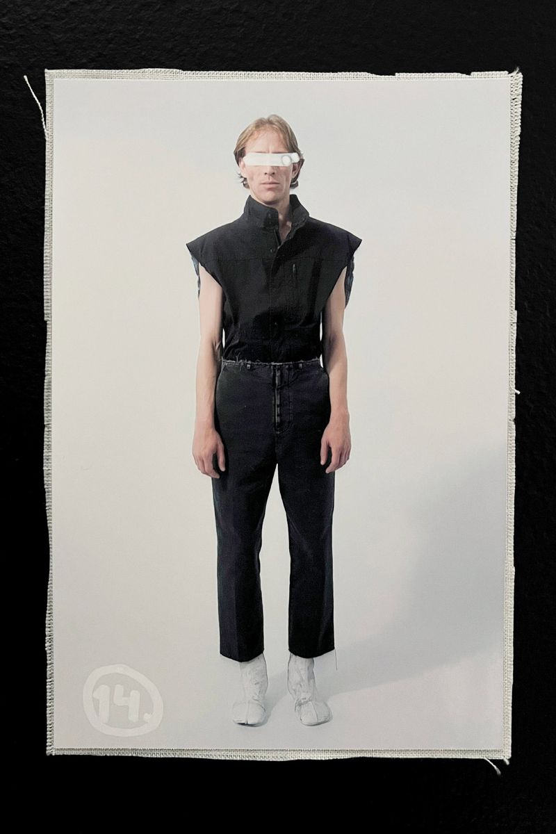 Adult male fashion model in black sleeveless top and black pants against a light background with white eyewear and white boots.