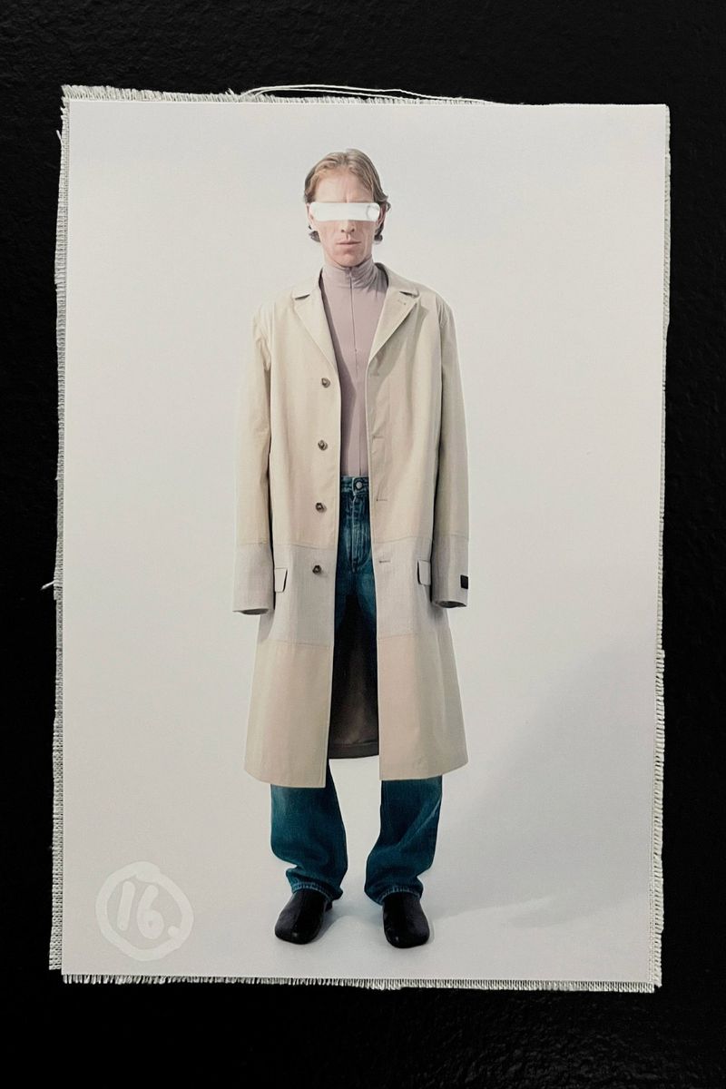 Full-length fashion portrait of a blond adult in a beige trench coat, lavender turtleneck, and jeans against a plain white backdrop.