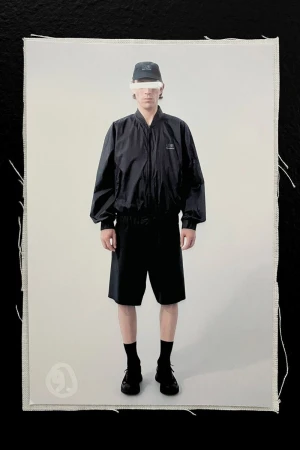 A person stands against a light grey backdrop wearing black athletic apparel, a cap, and shoes, presented on a rough canvas background.