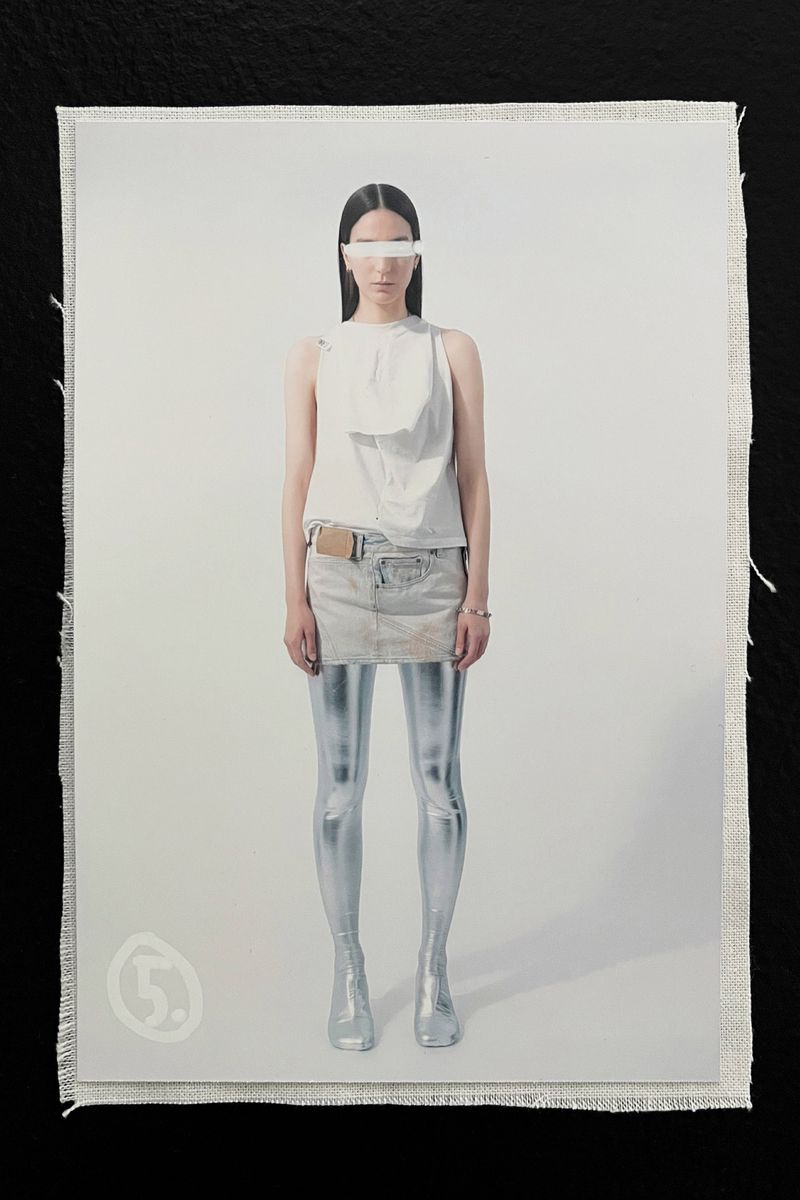 Fashion shot featuring a woman in white top and skirt, silver tights and boots, styled with a white band across her eyes, neutral backdrop.