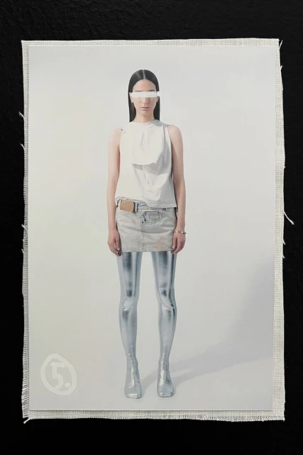 Fashion shot featuring a woman in white top and skirt, silver tights and boots, styled with a white band across her eyes, neutral backdrop.